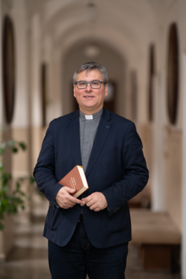 Docent Jozef Jančovič v tmavomodrom saku s Bibliou v ruke stojí na chodbe.